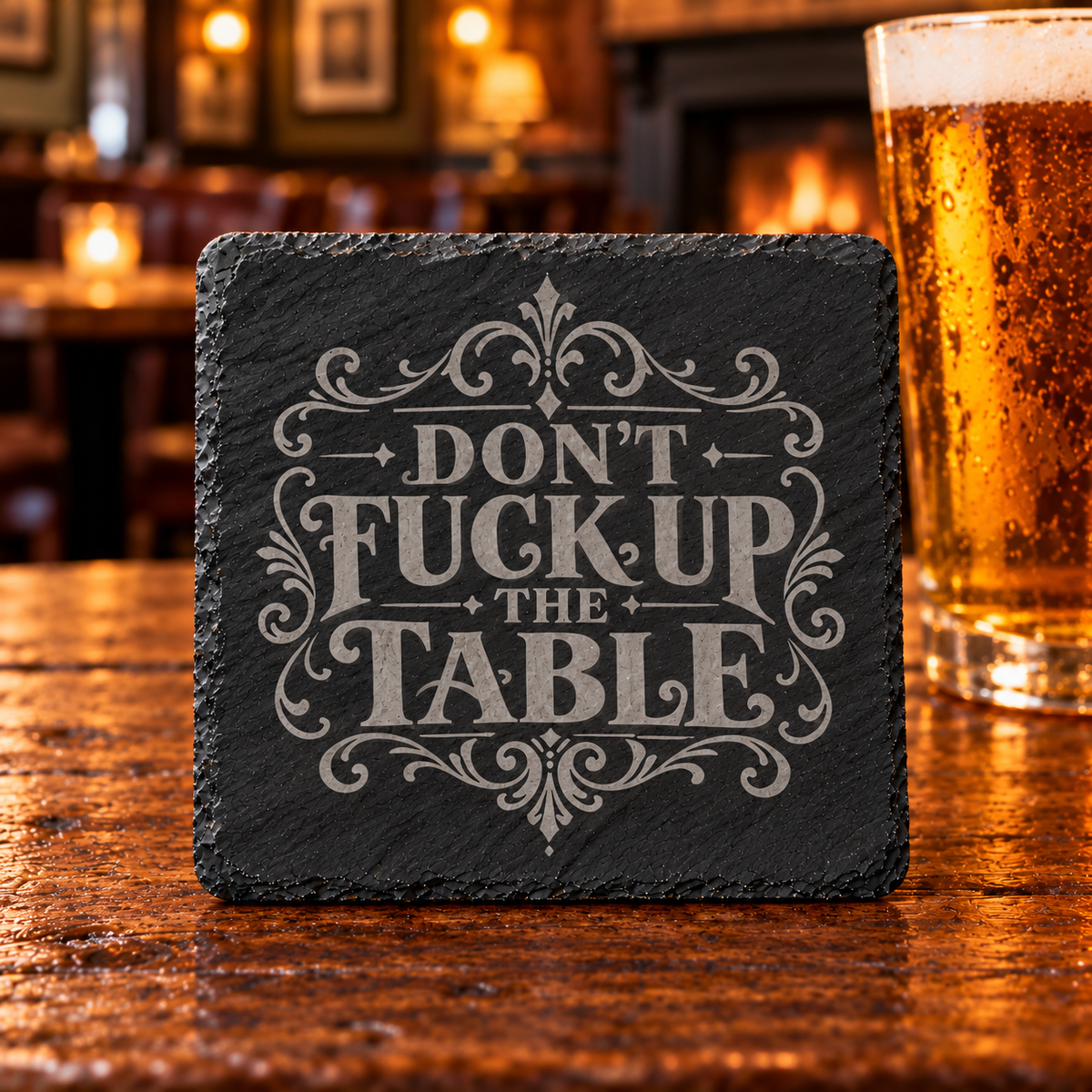 Fyrkantigt glasunderlägg i skiffer med texten Don’t Fuck Up the Table och dekorativa ornament, fotograferat uppifrån.