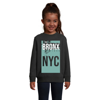 Barn i svart sweatshirt med ett dämpat turkost rektangulärt motiv, vit Bronx-text, svarta NYC-bokstäver och årtalet 1898.